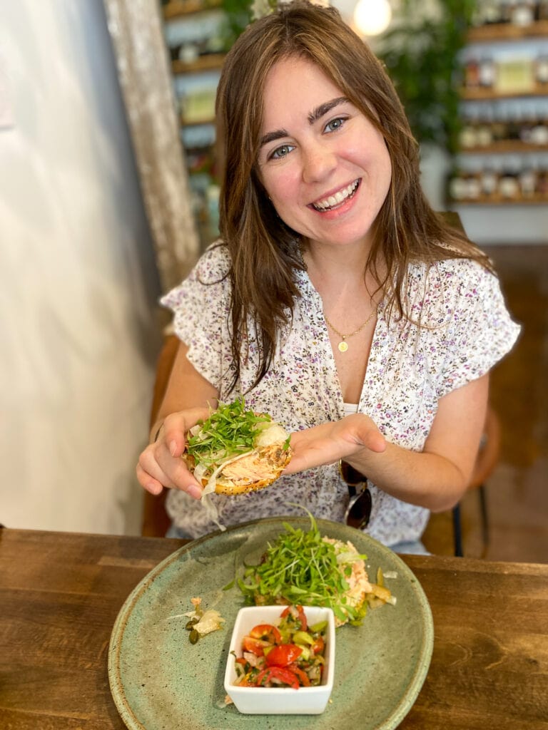 Sarah, a white woman with brown hair, smiles and holds a gluten free bagel.