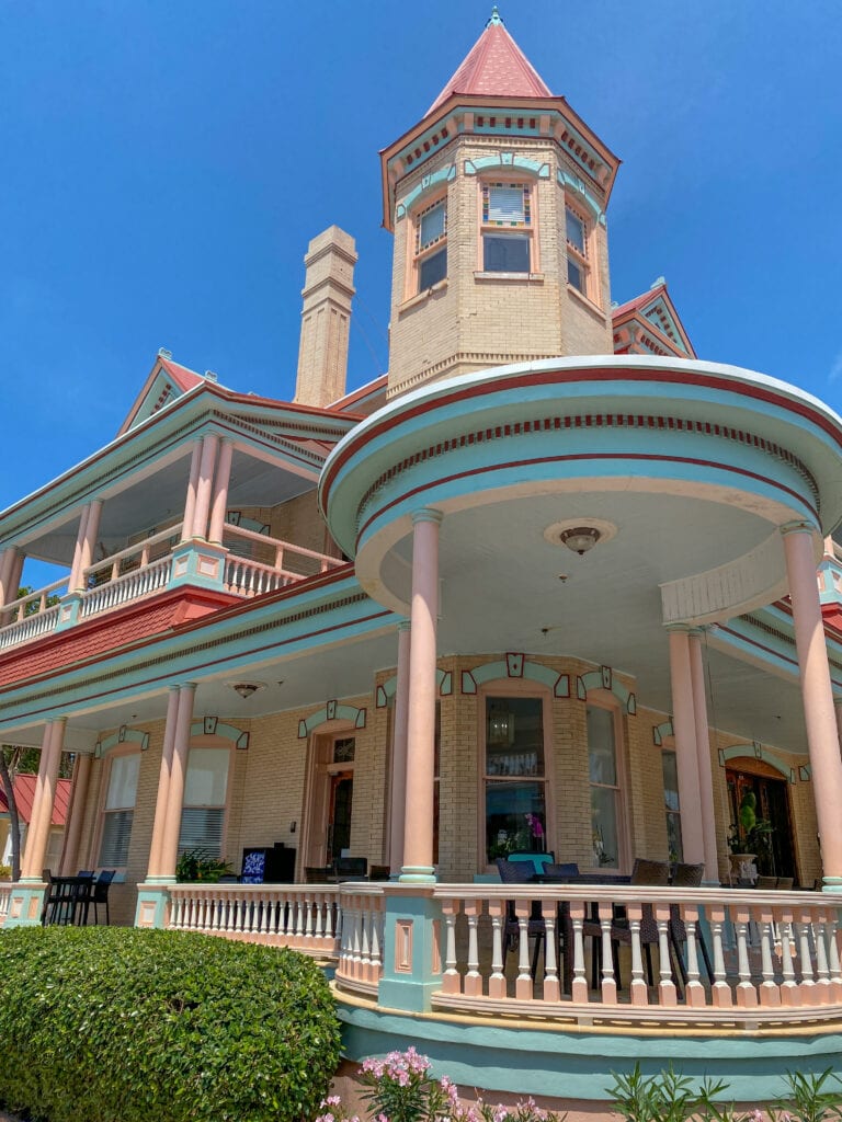 the southernmost mansion is light pink, white, and blue with a turret and wrap around porch and blue skies behind it.