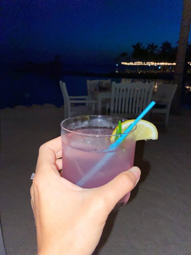 Sarah's hand holds a pink cocktail with a dark ocean in the background.