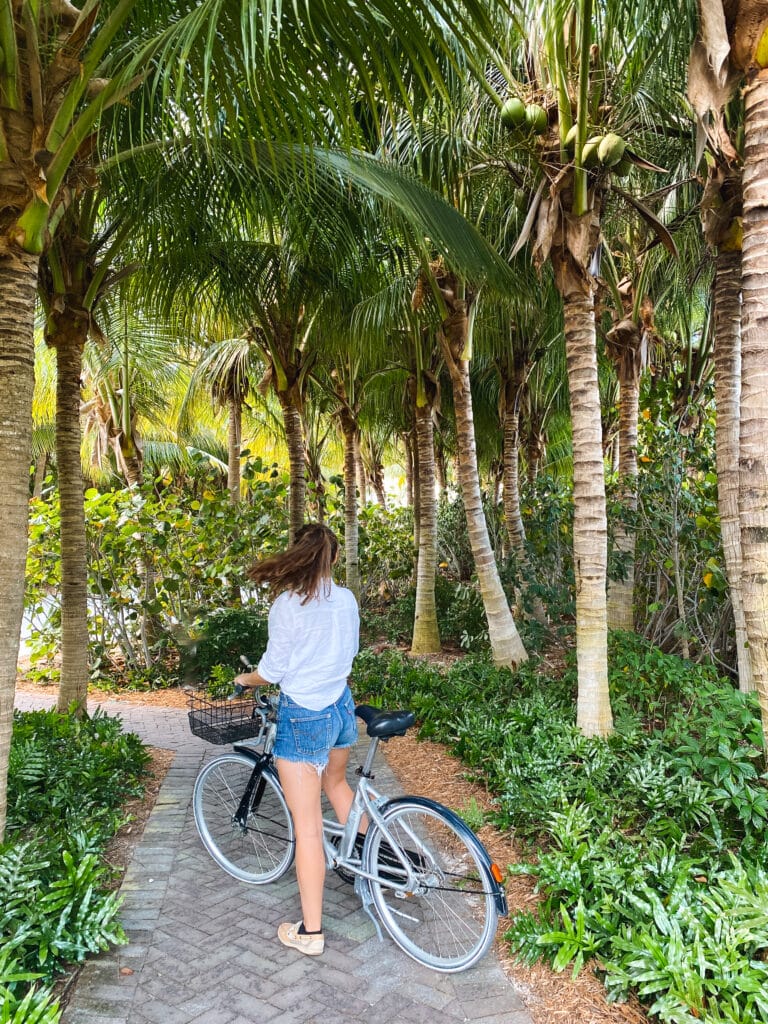 Sarah stands by a bike. She is wearing a white linen shirt and jean shorts, and looks away from the camera. Surrounded by tall palm trees.