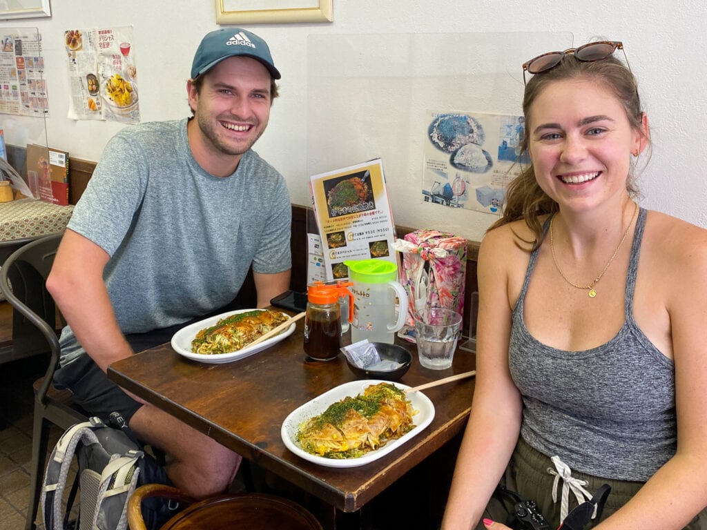 Sarah and Dan smile, sitting at table with hiroshima-style gluten free okonomiyaki. 