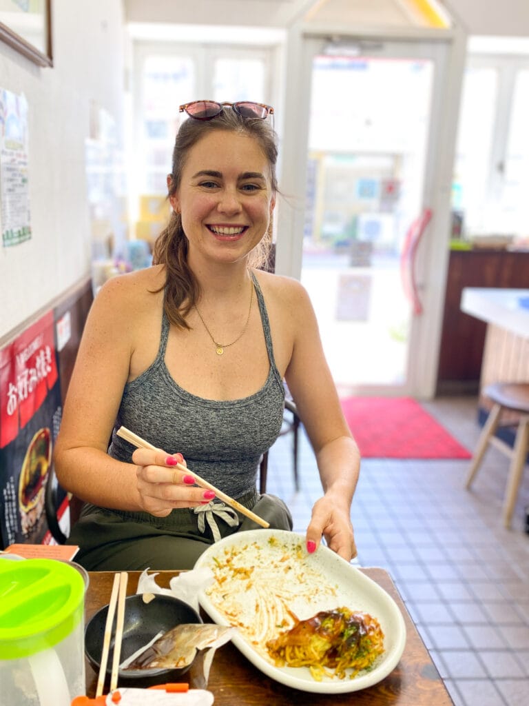 Sarah smiles, holding chopsticks and eating gluten free okonomiyaki.