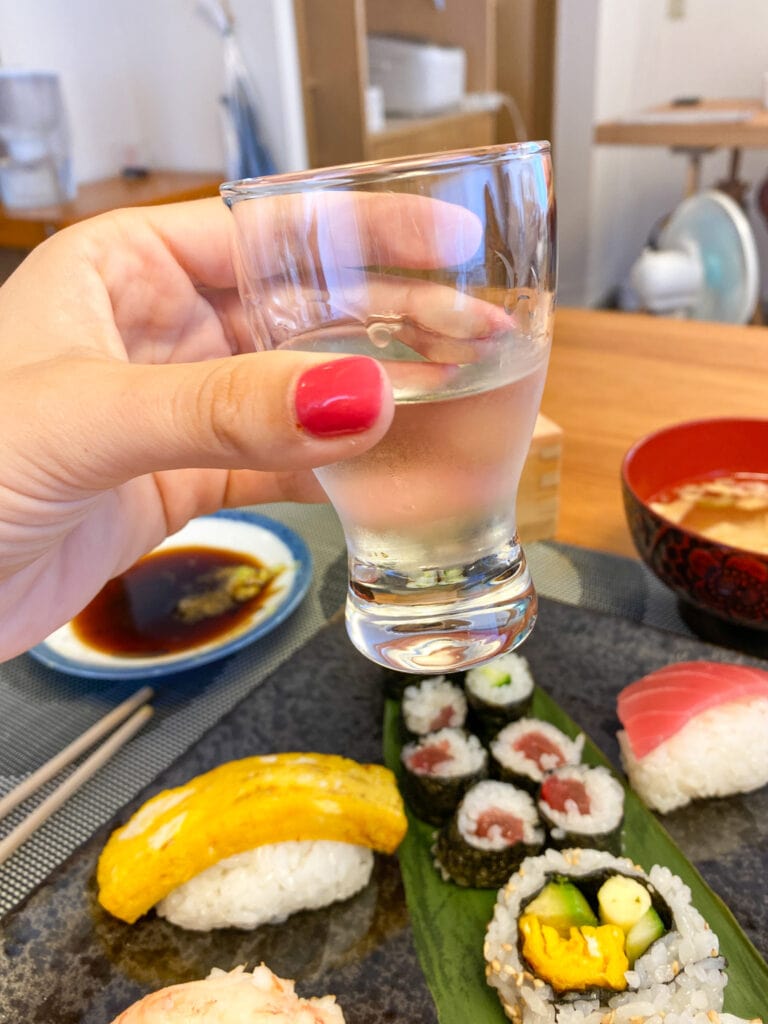Sarah's hand holding glass of sake in front of sushi plate.