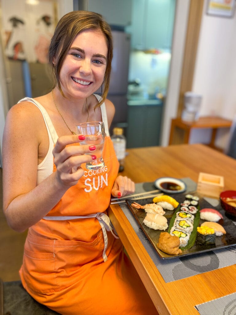 Sarah smiles and holds sake next to plate of sushi made in class.