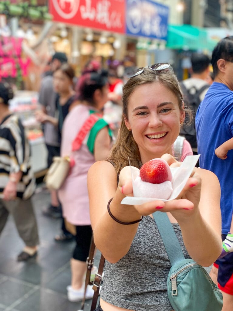 Sarah smiles and holds a strawberry daifuku at Kuromon Ichiba Market in Osaka Japan.