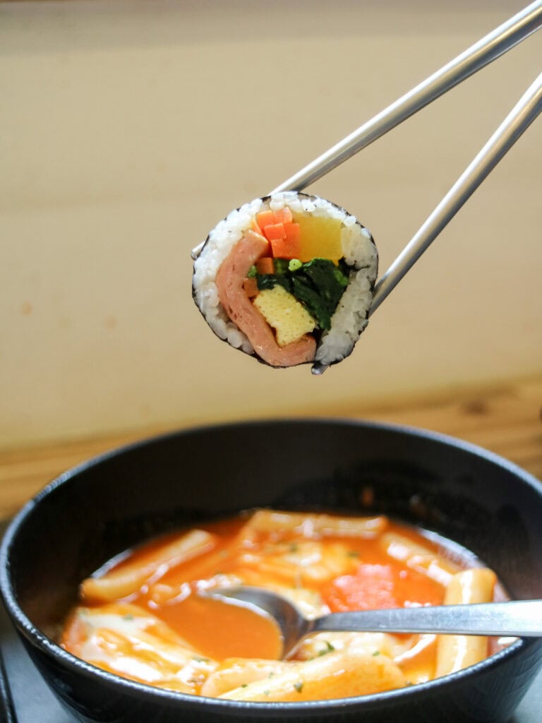 Chopsticks holding gimbap with tteok-bokki in the background