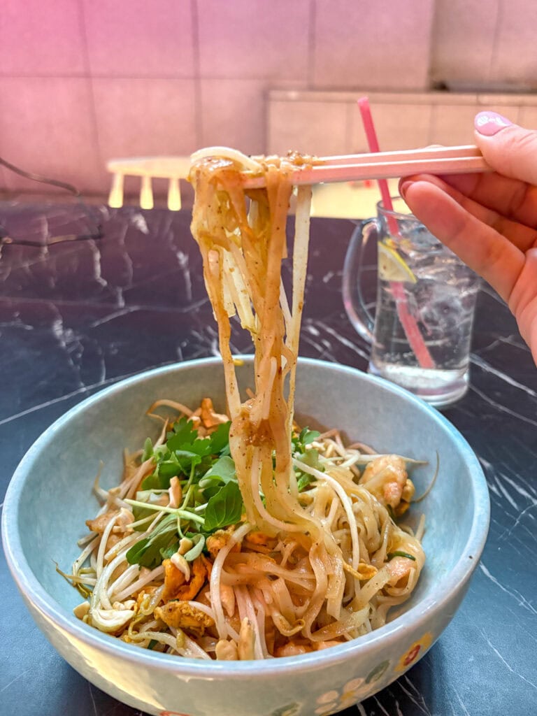 Bowl of Asian noodle salad with vegetables and herbs, chopsticks lifting noodles.