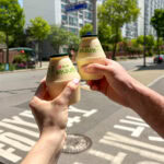 Two hands holding bottled banana milk drinks outdoors in an urban setting in South Korea.
