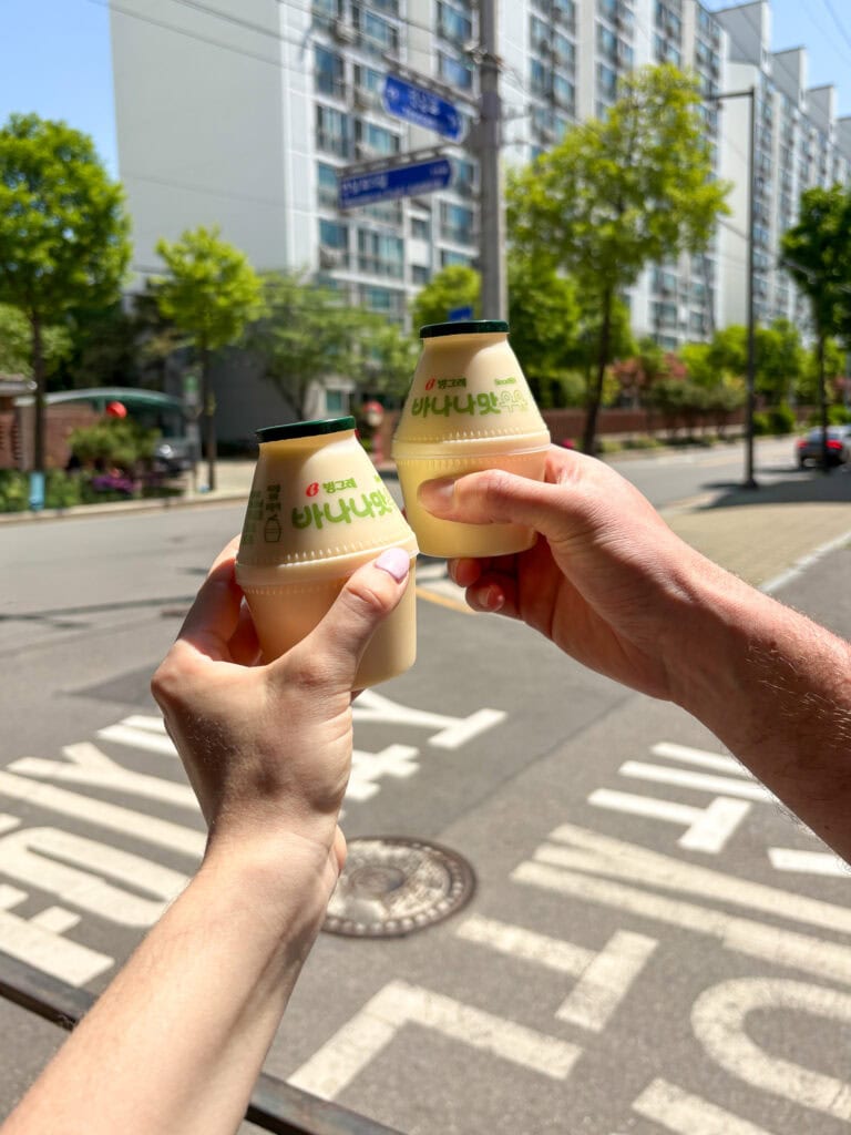 Two hands holding bottled banana milk drinks outdoors in an urban setting in South Korea.