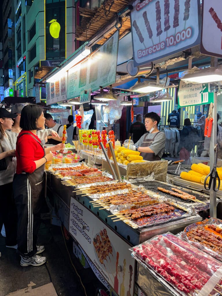 Vibrant street food stall in South Korea with grilled meats and corn at night.