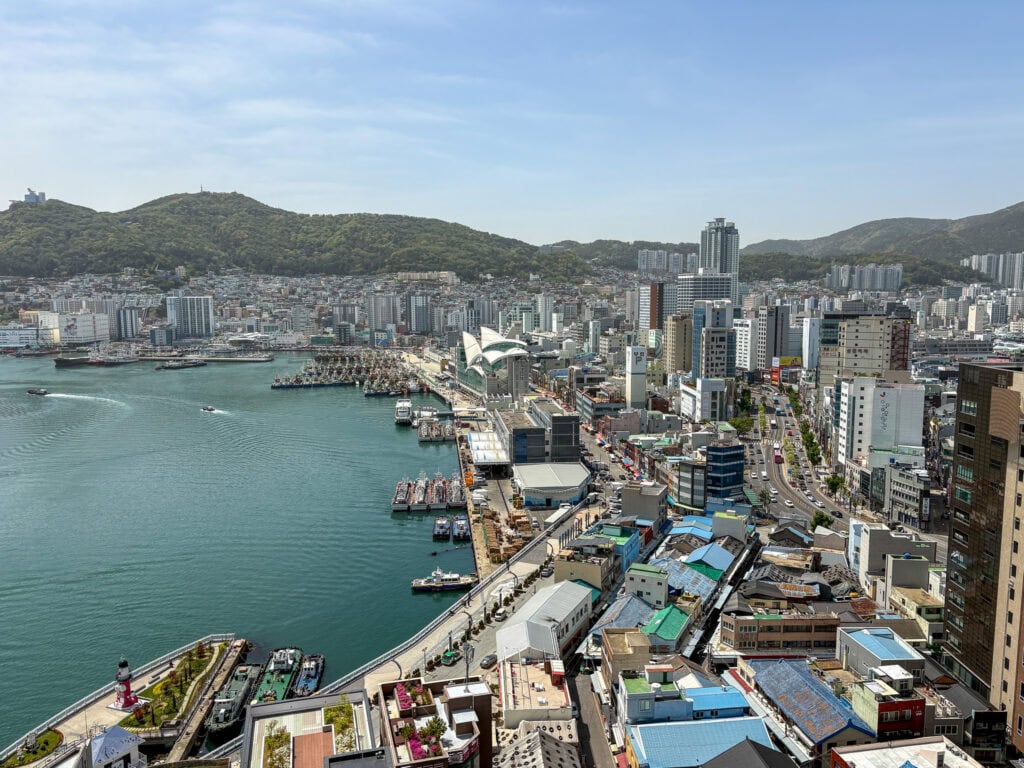 Panoramic view of Busan's skyline and harbor with mountains in the background.