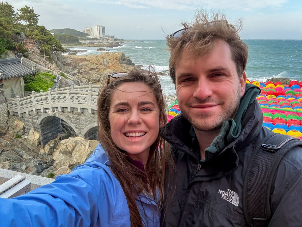 Happy couple taking a selfie at Busan's rocky coastline and colorful beach umbrellas.