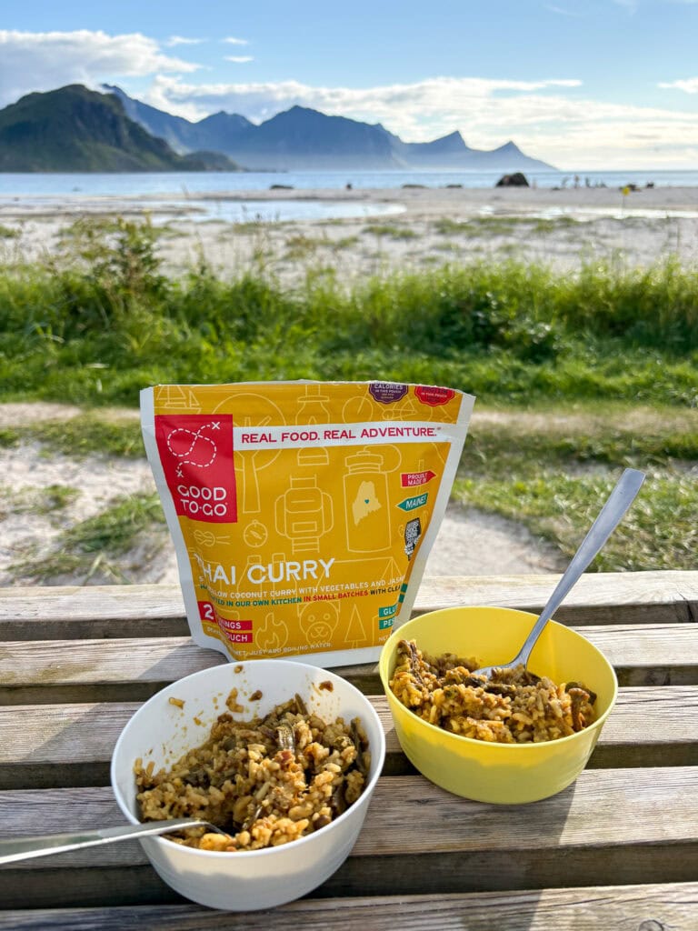 Outdoor meal with curry on a wooden table near a beach with mountains in the background.