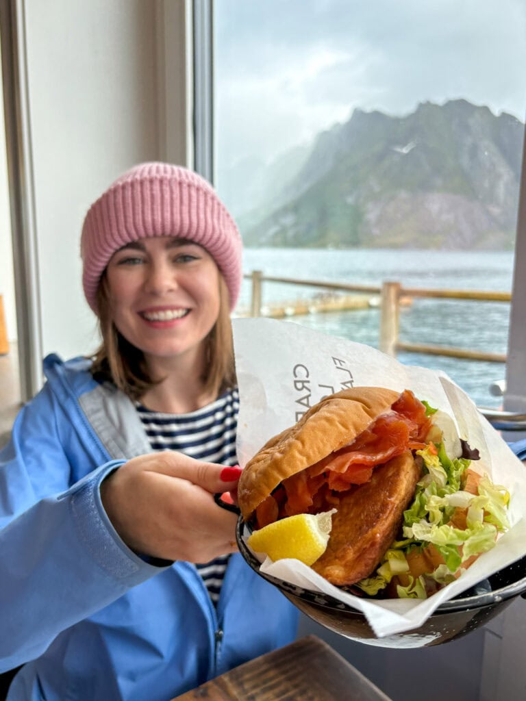 Smiling woman holding a delicious fish sandwich with scenic mountain view.