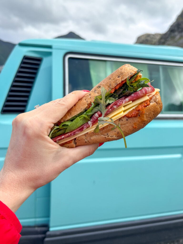 Hand holding a gluten-free sandwich with fresh vegetables in front of a blue van.