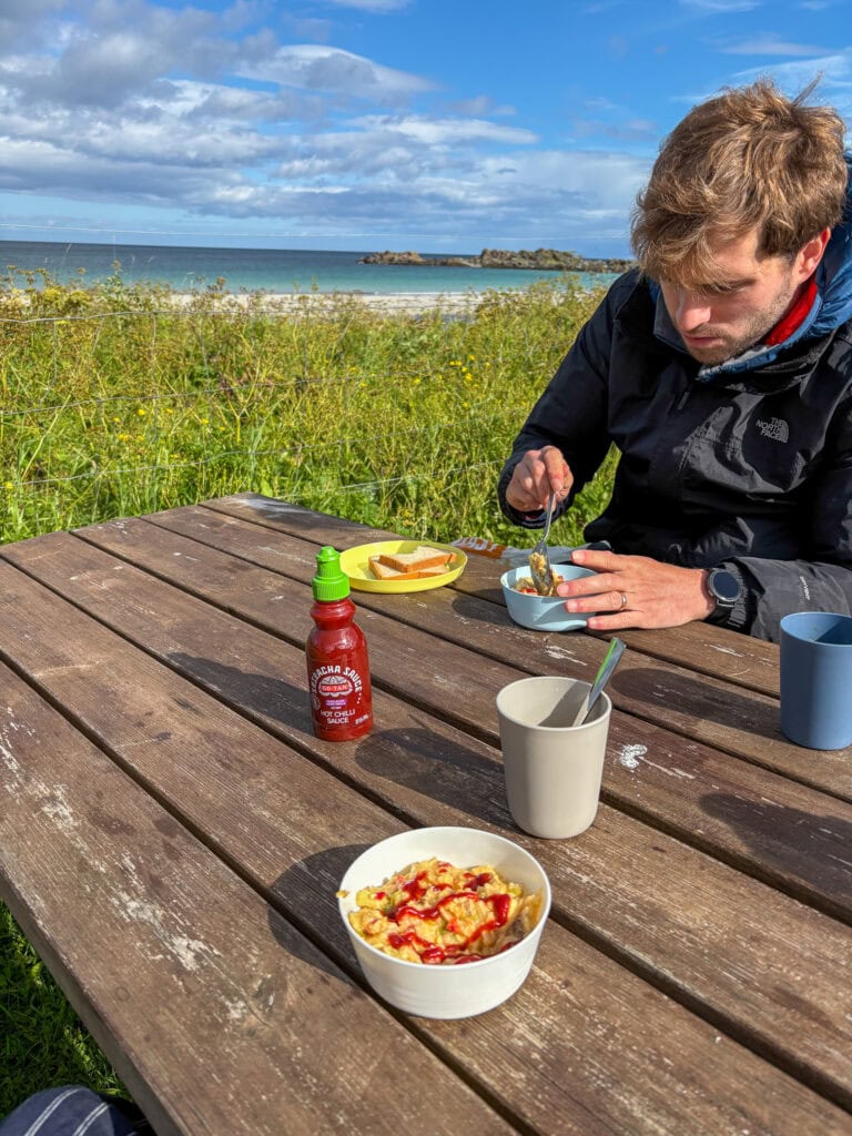 Man enjoying a meal outdoors on a wooden table near the coast with a bowl of food and condiments.