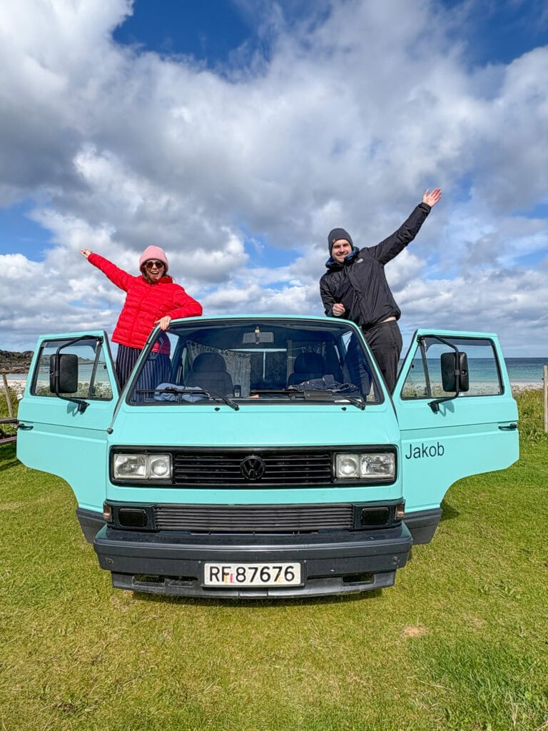 Two people standing on a light blue van with open doors outdoors under cloudy sky.