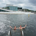People swimming in Oslo waterfront with modern architecture and Ferris wheel in background.
