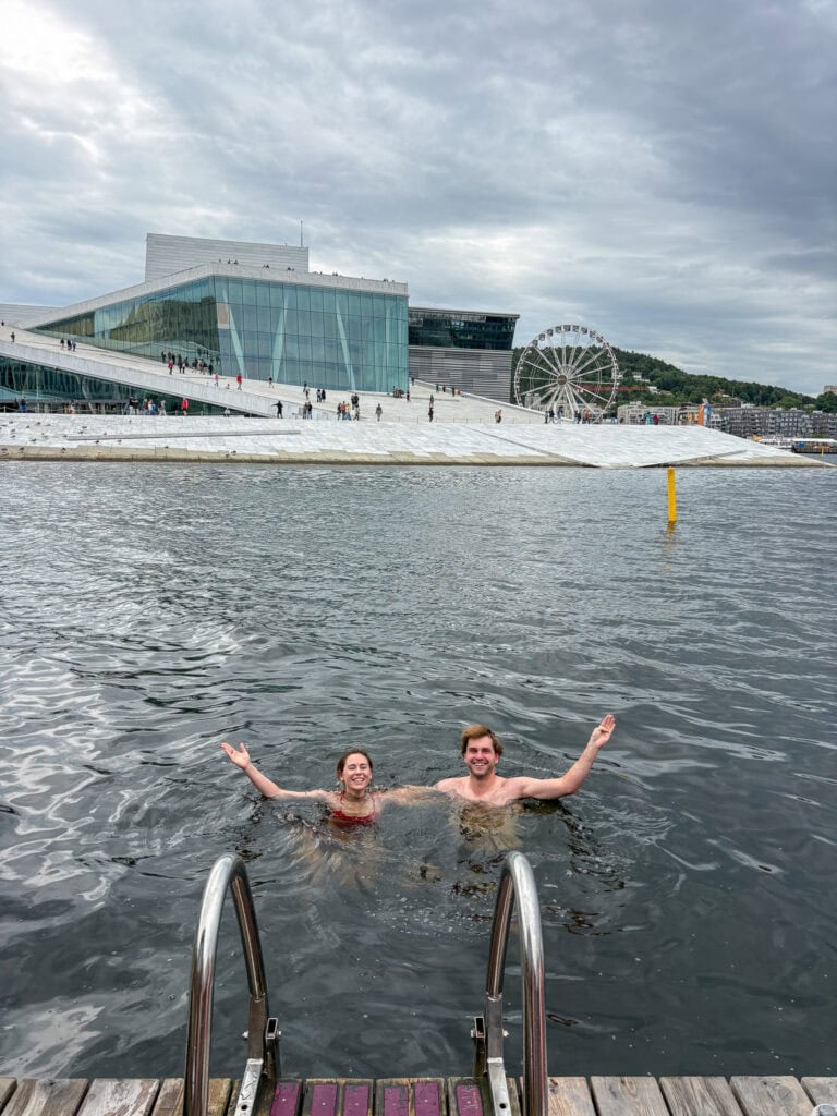 People swimming in Oslo waterfront with modern architecture and Ferris wheel in background.