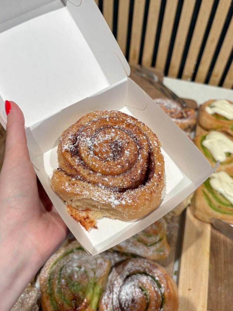 Fresh cinnamon roll with sugar in a white box on a hand, bakery display in background.