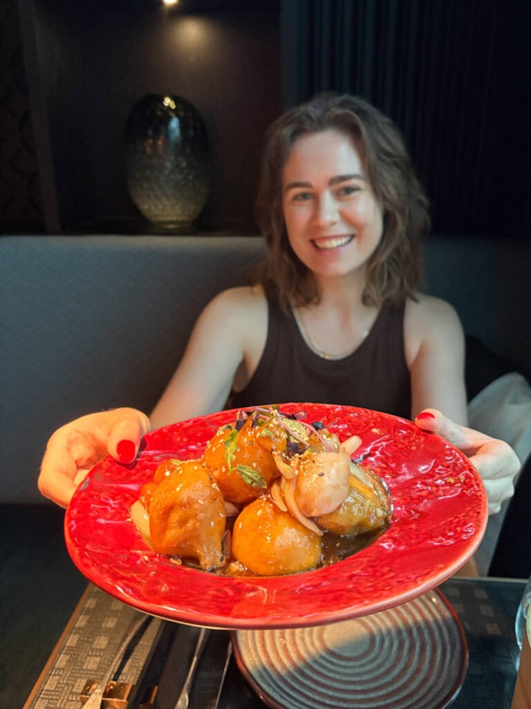 Smiling woman holding a red plate of glazed meatballs in a restaurant setting.