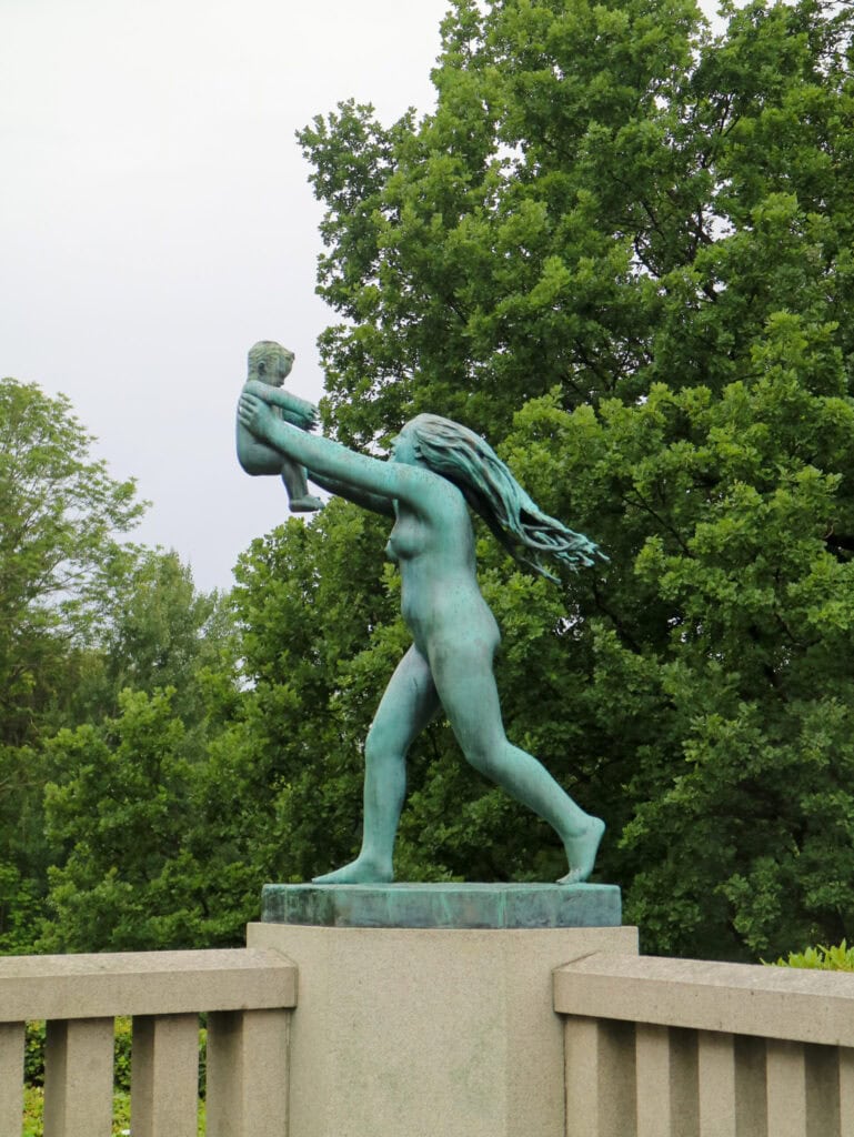 Bronze statue of a woman holding a child high above her head in a park setting.