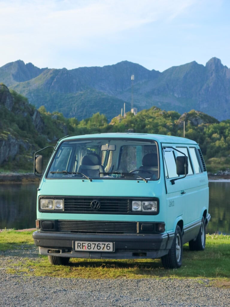 Blue Volkswagen van parked near a lake with mountains in the background.