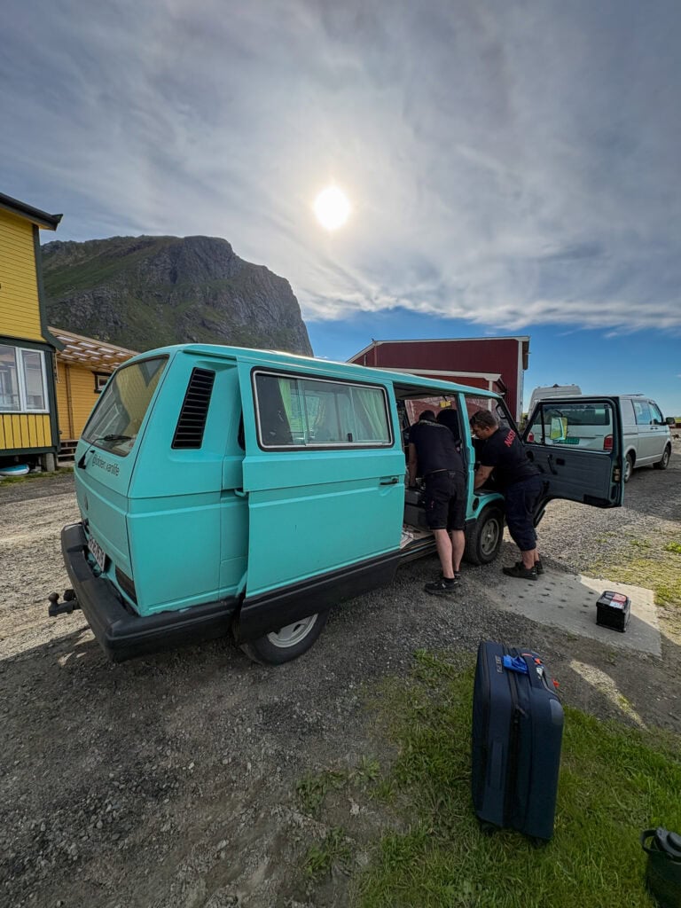 Vintage teal camper van parked outdoors with people loading luggage, scenic mountain background.