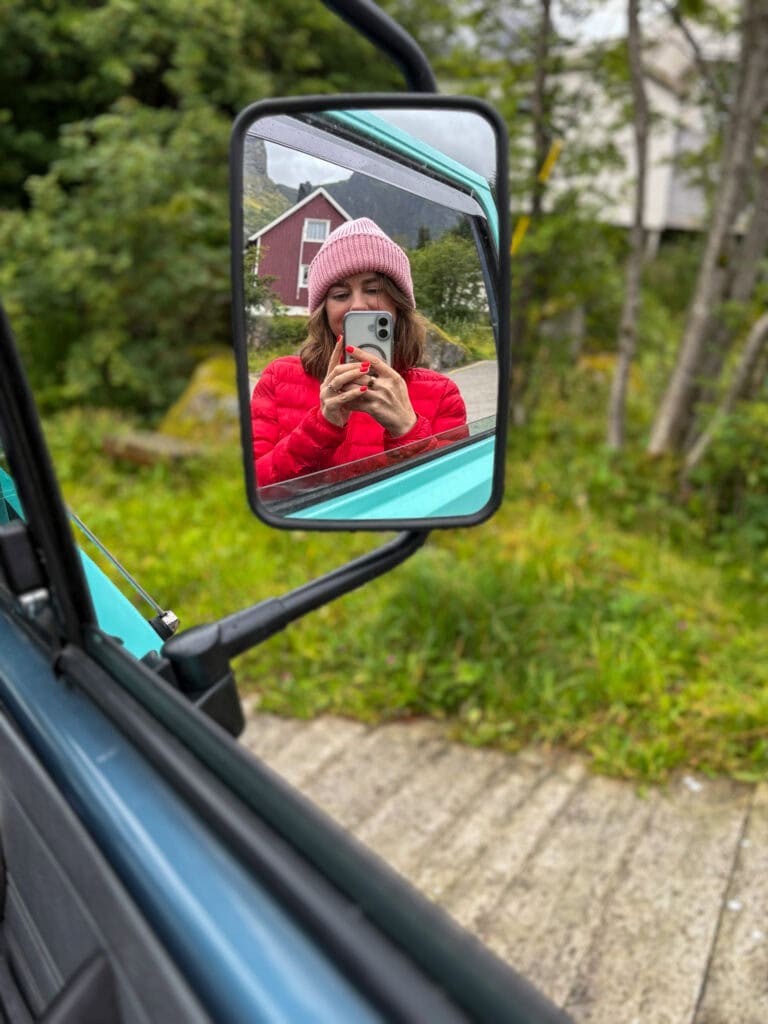Woman in pink hat taking a selfie reflected in a vehicle's side mirror.