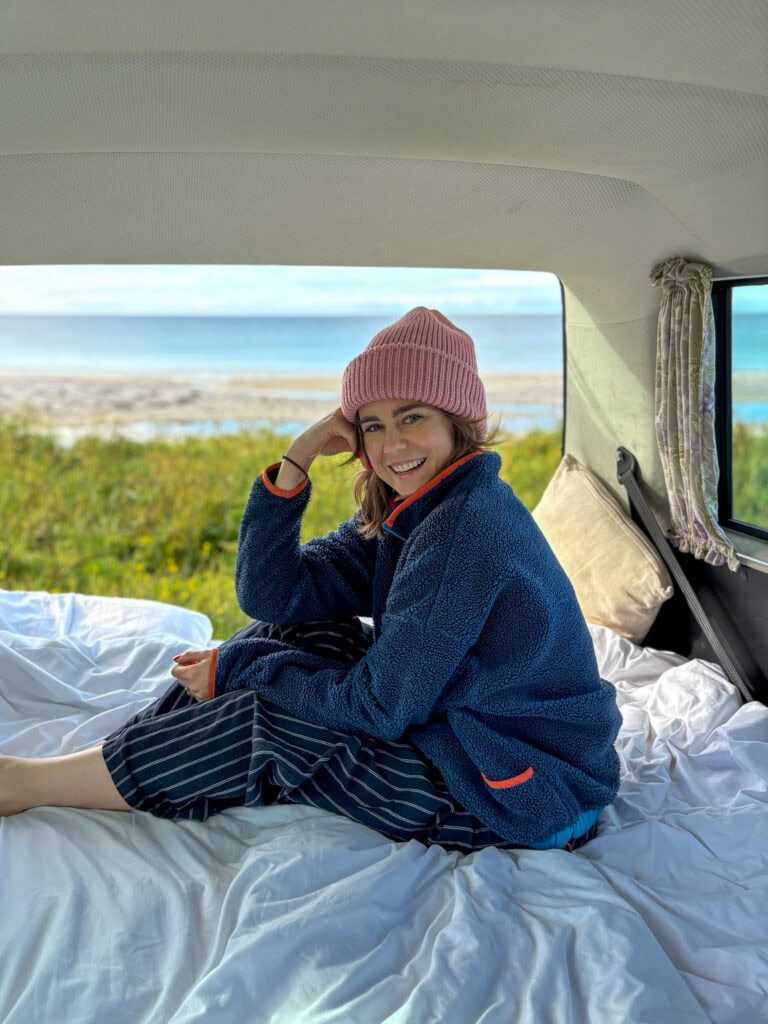 Woman in cozy fleece jacket and pink beanie inside camper van by the sea.