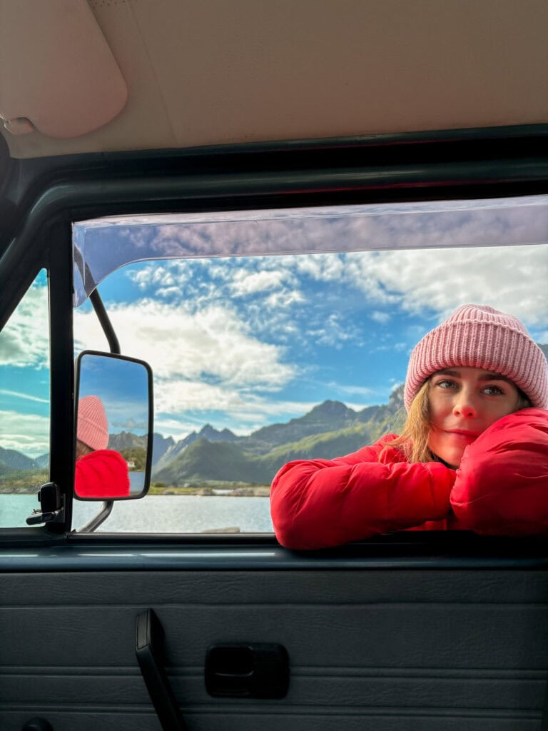 Woman in pink beanie and red jacket looking out of van window at mountain landscape.