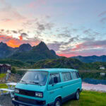 Blue Volkswagen camper van parked near a mountain lake at sunset.