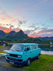 Blue Volkswagen camper van parked near a mountain lake at sunset.