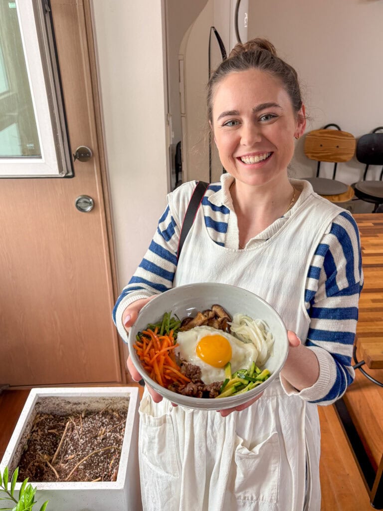 Smiling woman holding a bowl of Korean food in a cooking class setting.