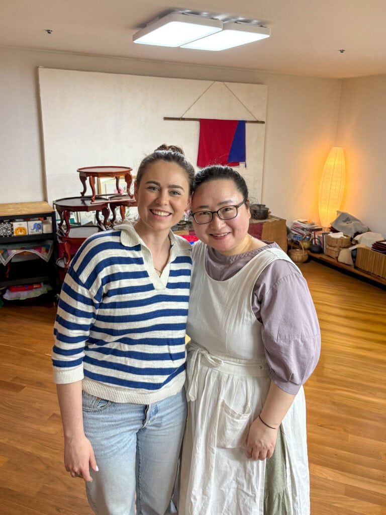 Two women smiling in a cozy, well-lit room with wooden floors and traditional decor.