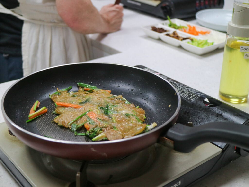 Cooking a Korean pancake on a stovetop with fresh ingredients nearby.