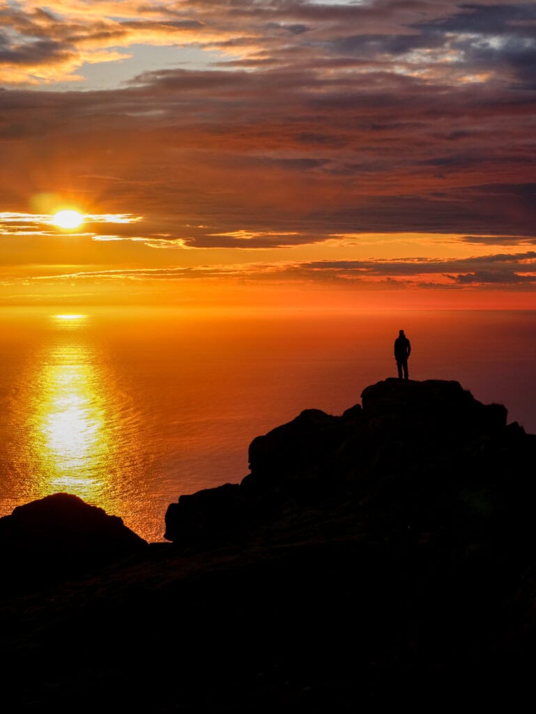 Person standing on rocks during a vibrant sunset over the ocean.
