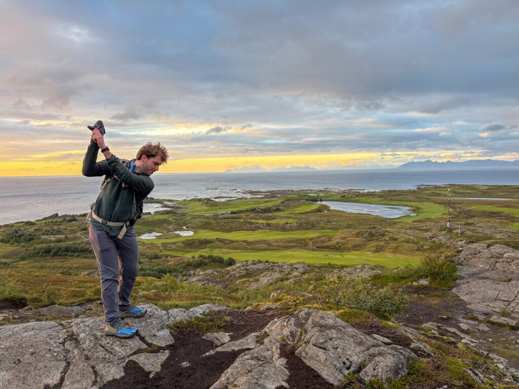 Young man playing golf on a rocky cliff overlooking Lofoten landscape at sunset.