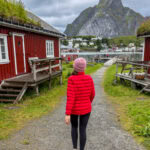 Woman in red jacket walking through Lofoten village with mountains in background.
