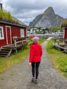 Woman in red jacket walking through Lofoten village with mountains in background.