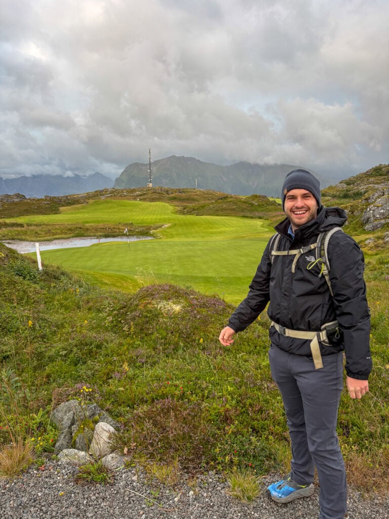 Hiker smiling on lush green landscape in Lofoten, Norway, with mountains and cloudy sky.